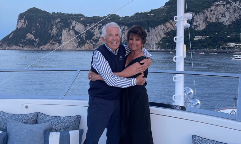 Man and woman embracing on the back deck of a boat with water and mountains in the background.