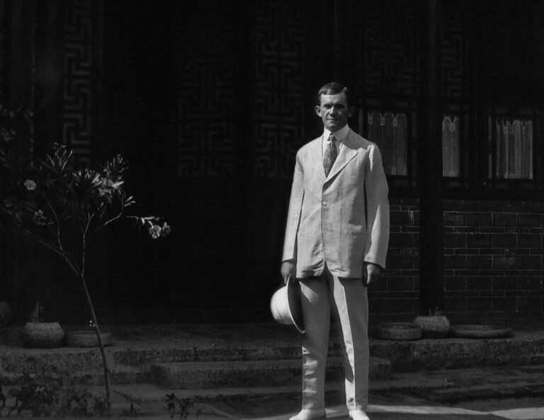 Man in suit holding a pith helmet in front of a building in the early 20th century.